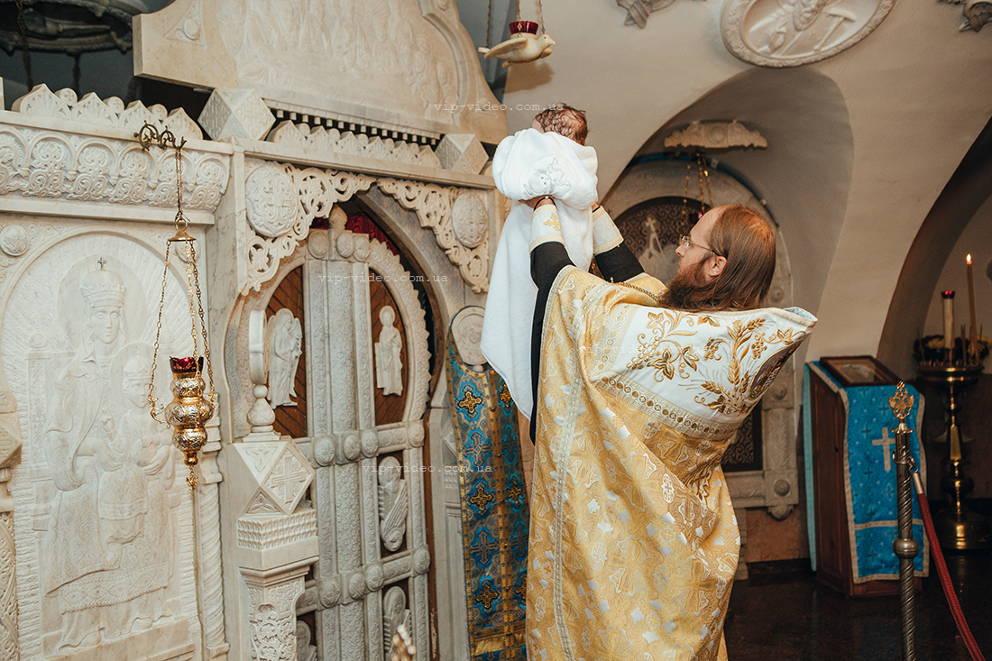 Фотографія на якій проходить обряд Хрещення диттину в церкві, а саме священник, руками піднятими до гори тримає малюка на фоню церковного Алтарю